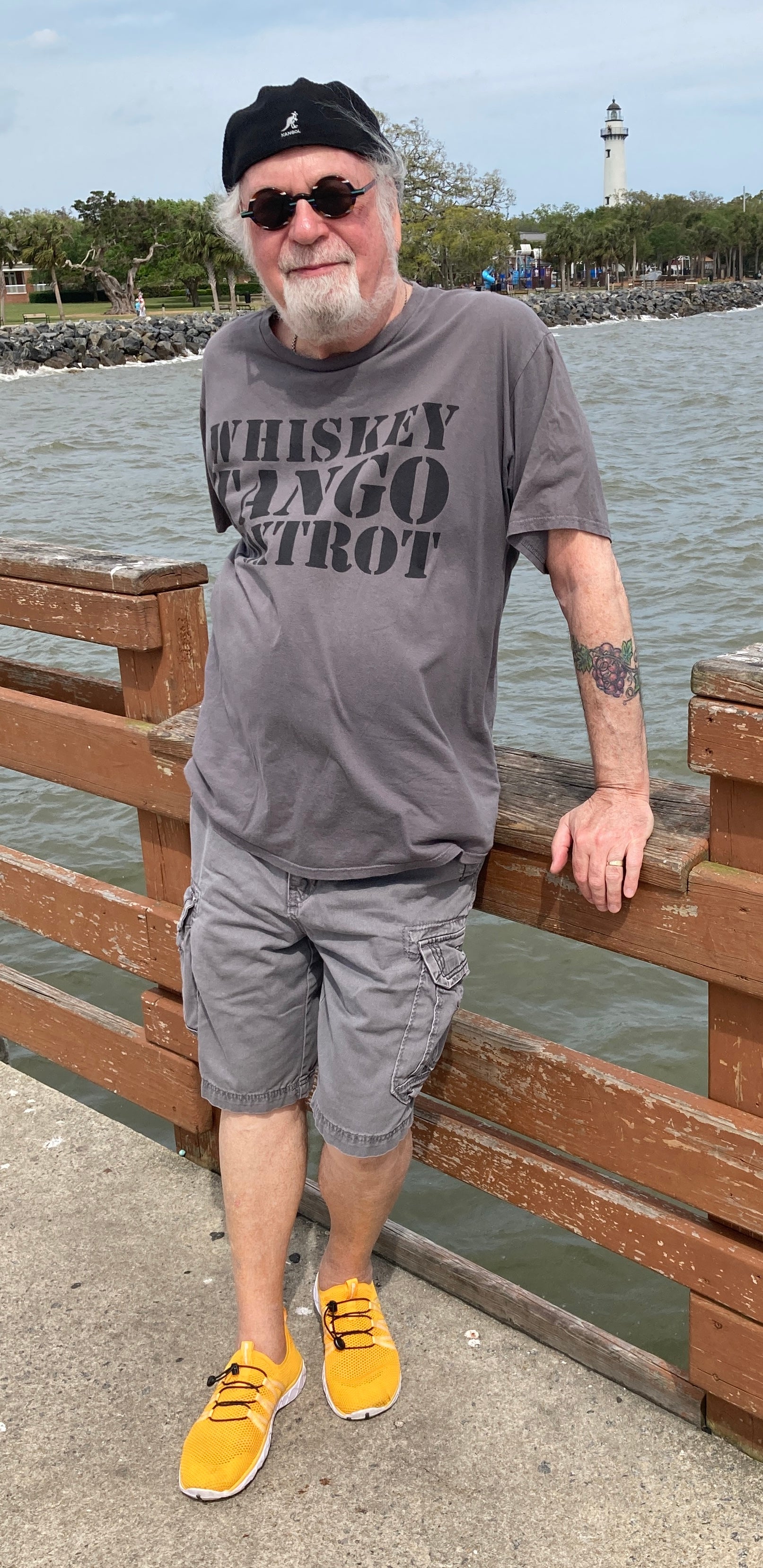 Man wearing a gray t-shirt with text, sunglasses, and yellow shoes standing on a wooden pier by water. Bill Citara's Artist Profile Picture